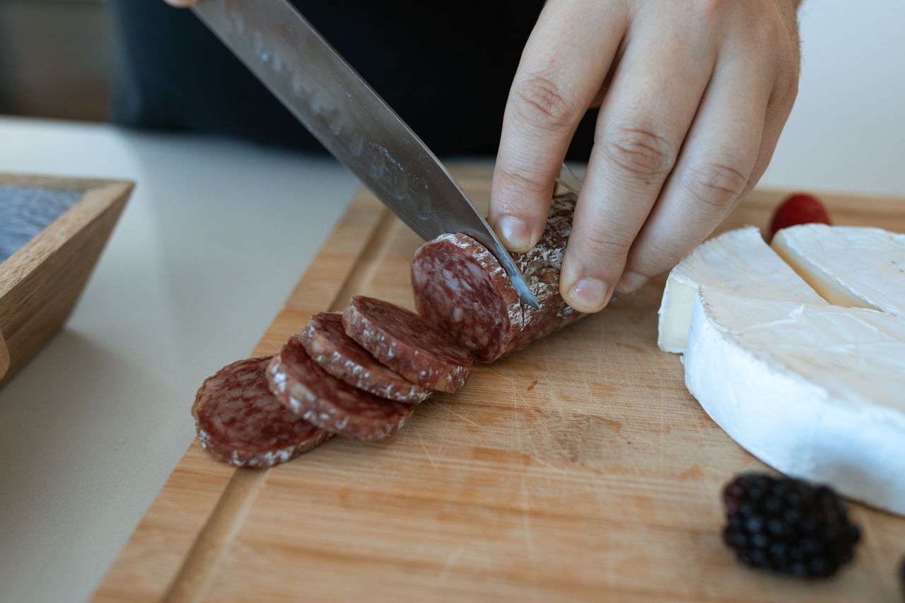 Hand slicing salami on wooden board with cheese and berries. Perfect for food concepts.